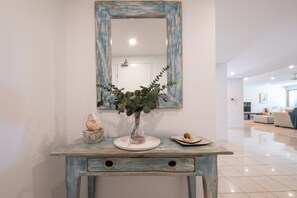 A room with a white wall, a blue-framed mirror, a small table with a vase of green plants, and a decorative shell.