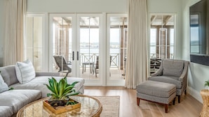 Amplia sala de estar con sofá, sillón y reposapiés, una mesa de centro con una planta, y vistas a una terraza exterior con sillas a través de puertas de cristal.