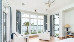 Una habitación de hotel moderna con dos sofás blancos frente a un amplio ventanal con vista a una masa de agua, una cama, una pequeña mesa redonda y una lámpara.