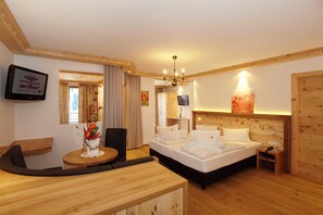 A hotel room with a wooden bed, a round wooden table with a vase of flowers, a TV mounted on the wall, and a wooden door. - Schwarzer Adler (Nauders)