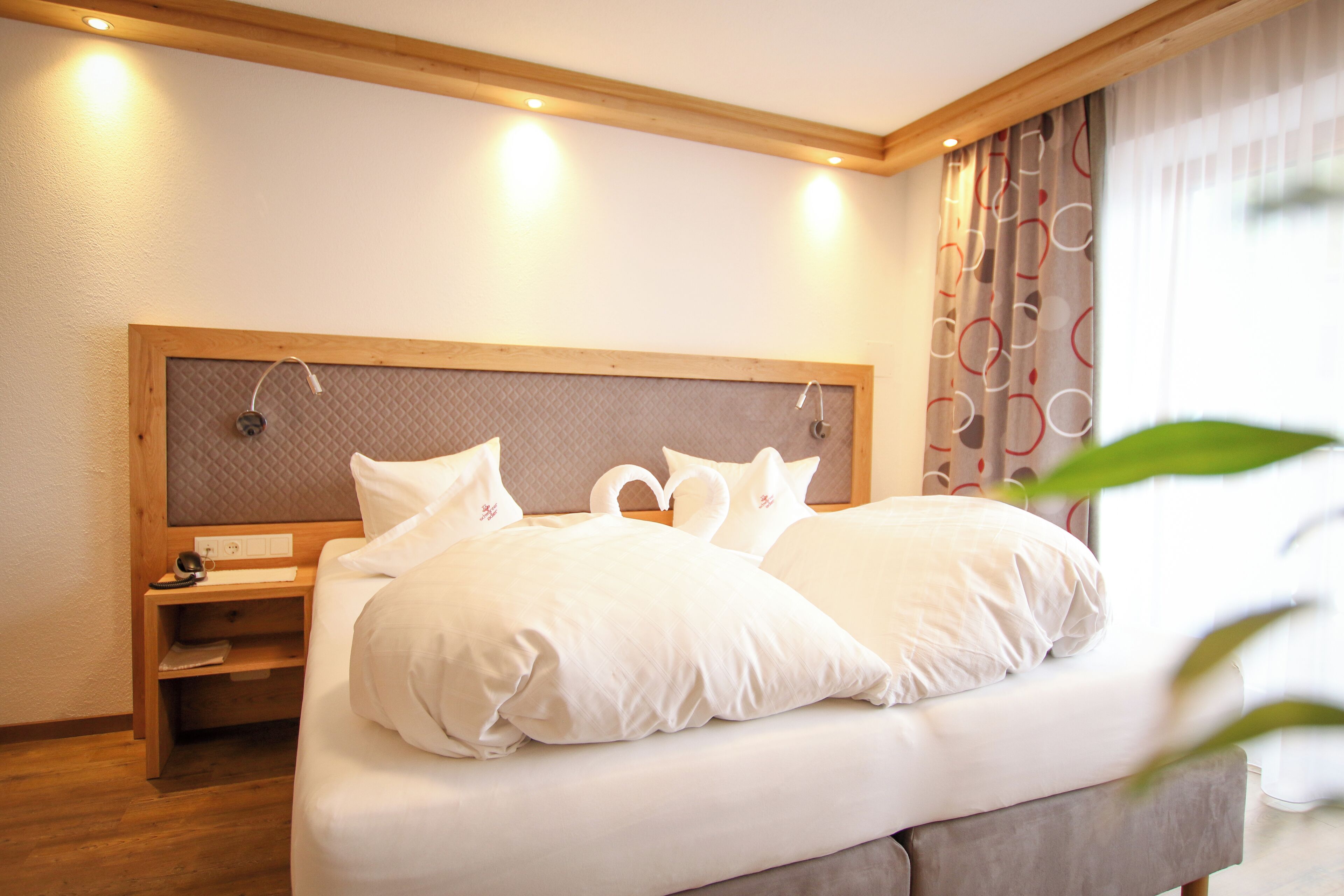 A neatly made bed with white linens and pillows, a wooden headboard, a bedside table with a lamp, and a window with curtains.