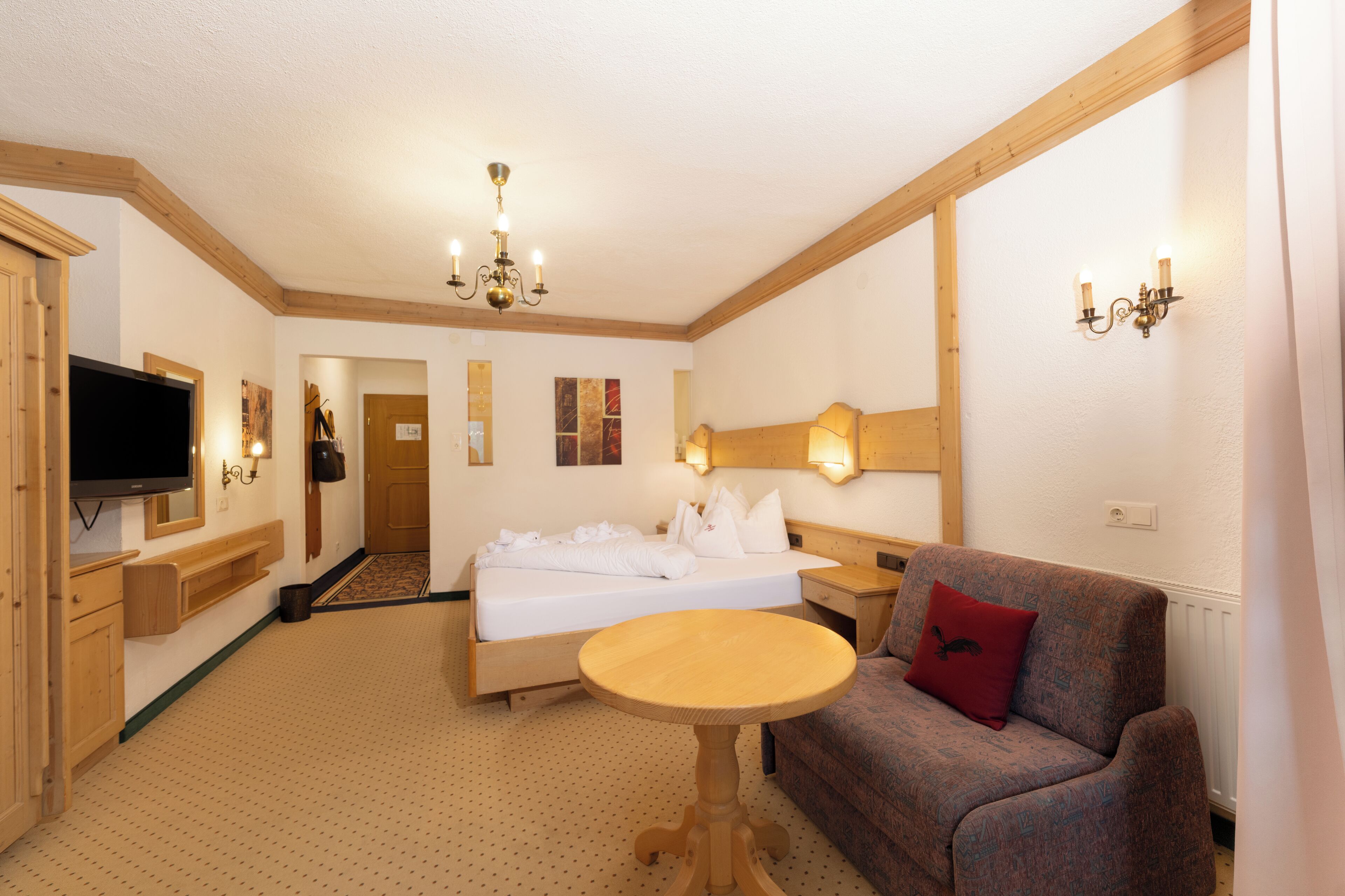A hotel room with a bed, a wooden table, a sofa, a television, and a chandelier.