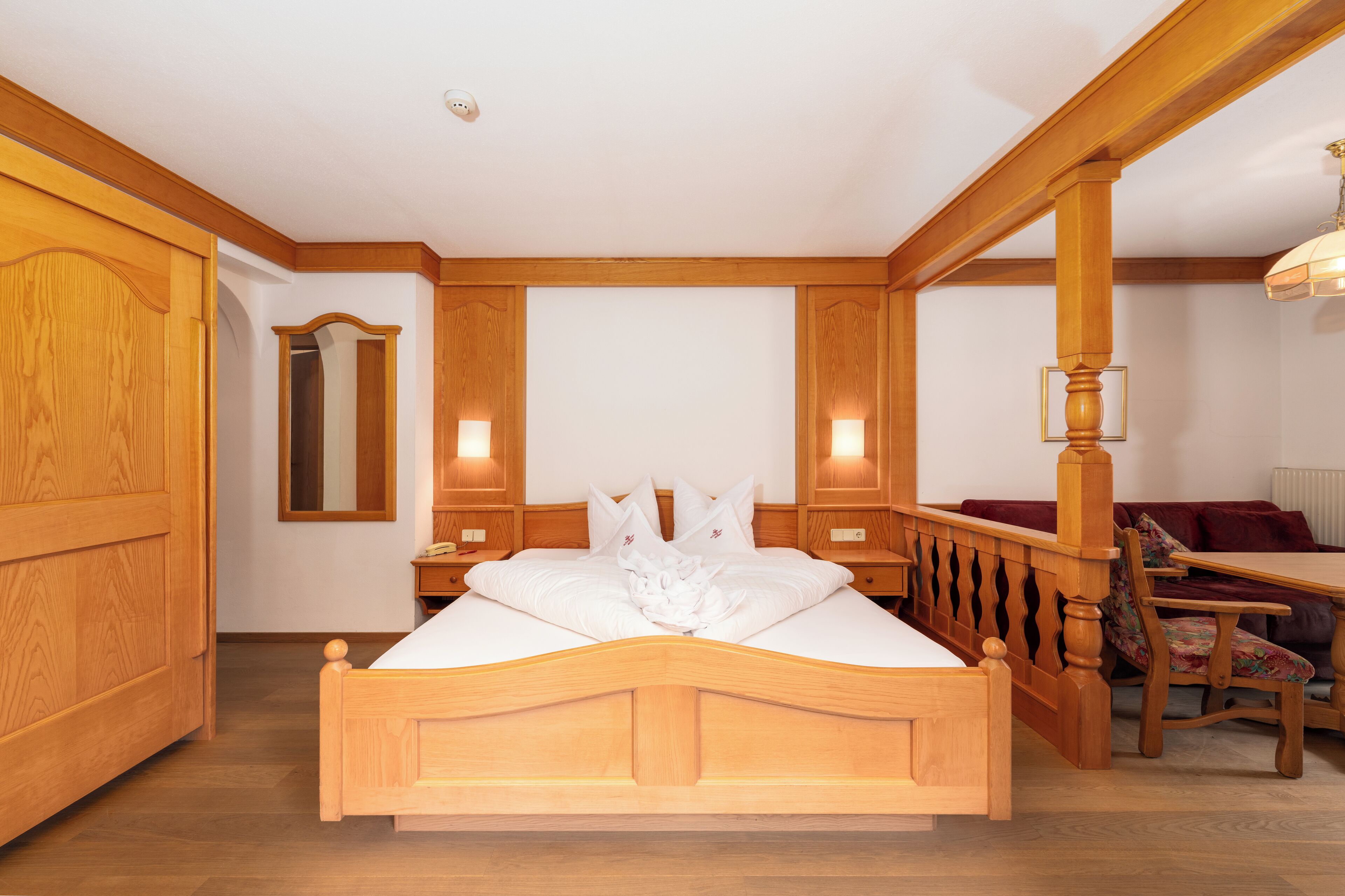 Une chambre à coucher avec un cadre en bois, comprenant un lit, des tables de chevet, un miroir et un coin salon avec une table et des chaises.