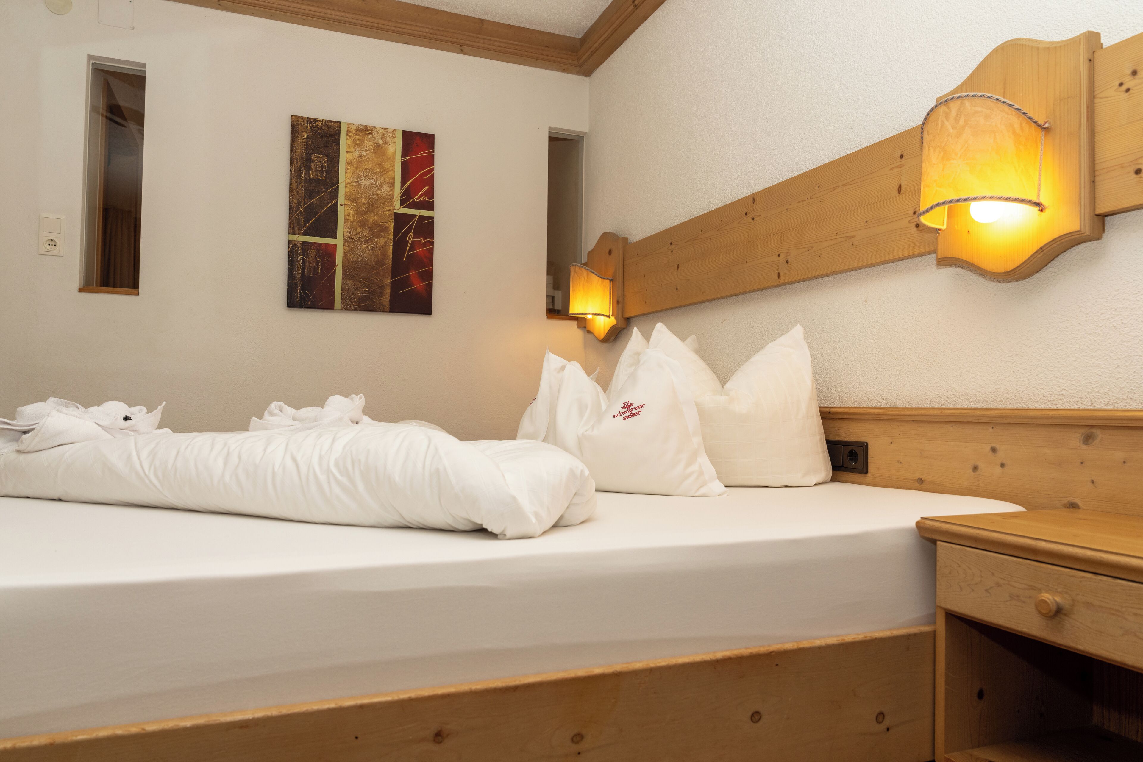 A neatly made bed with white linens and pillows, a wooden headboard, and a wall-mounted light fixture.