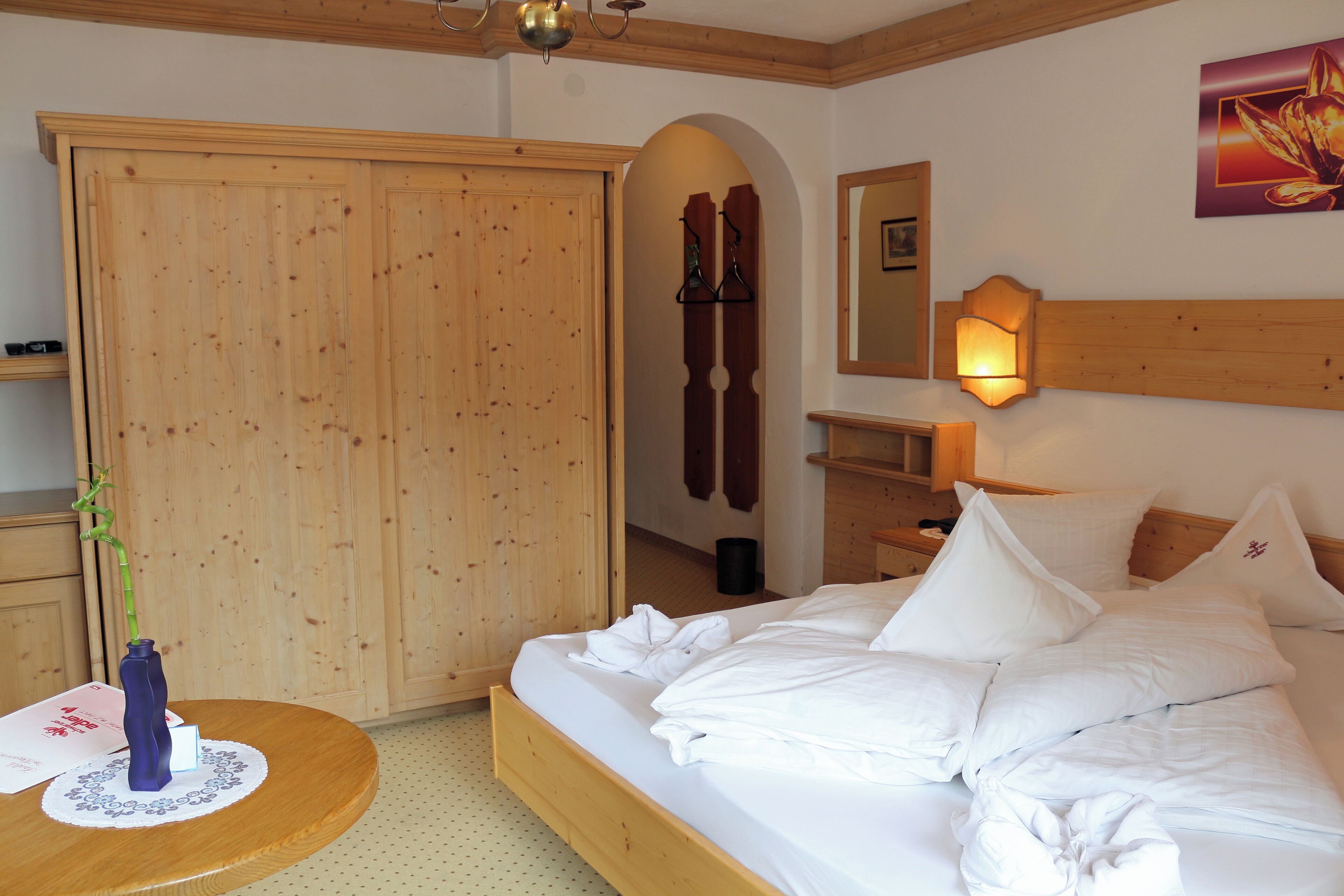 A hotel room with a wooden bed, white bedding, a round wooden table with a vase, and a wardrobe with a mirror.