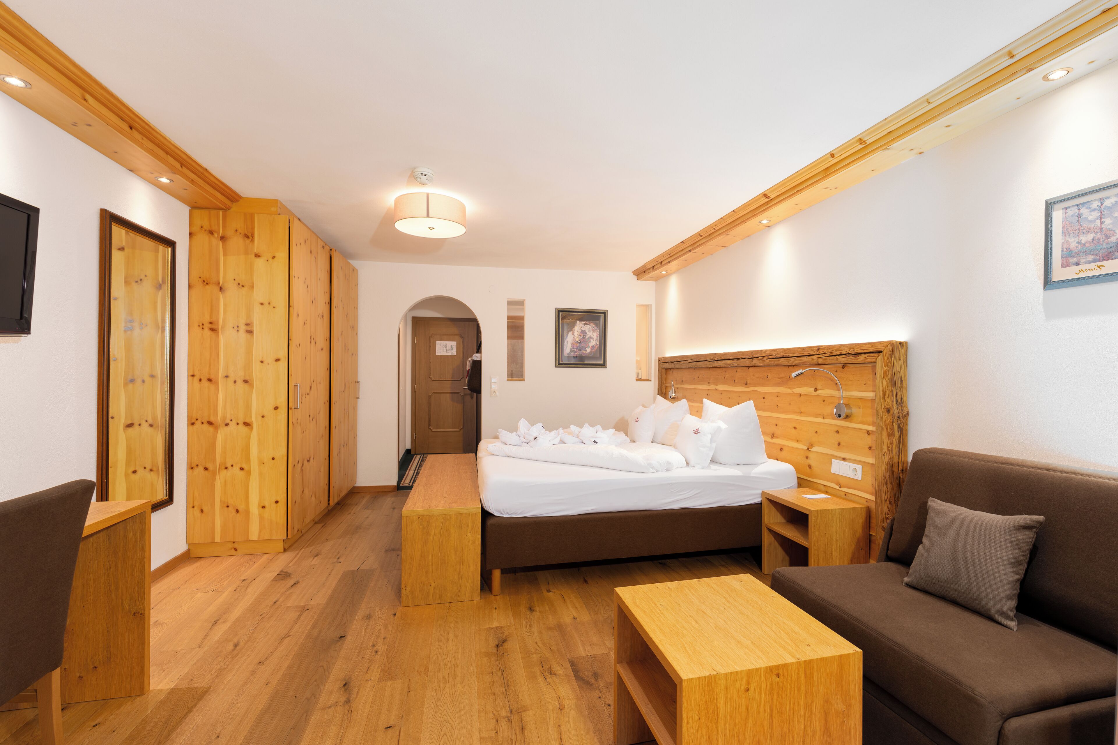 Una habitación de hotel con piso de madera, una cama con ropa blanca, un sofá marrón, una mesa de centro de madera y un cabecero de madera.