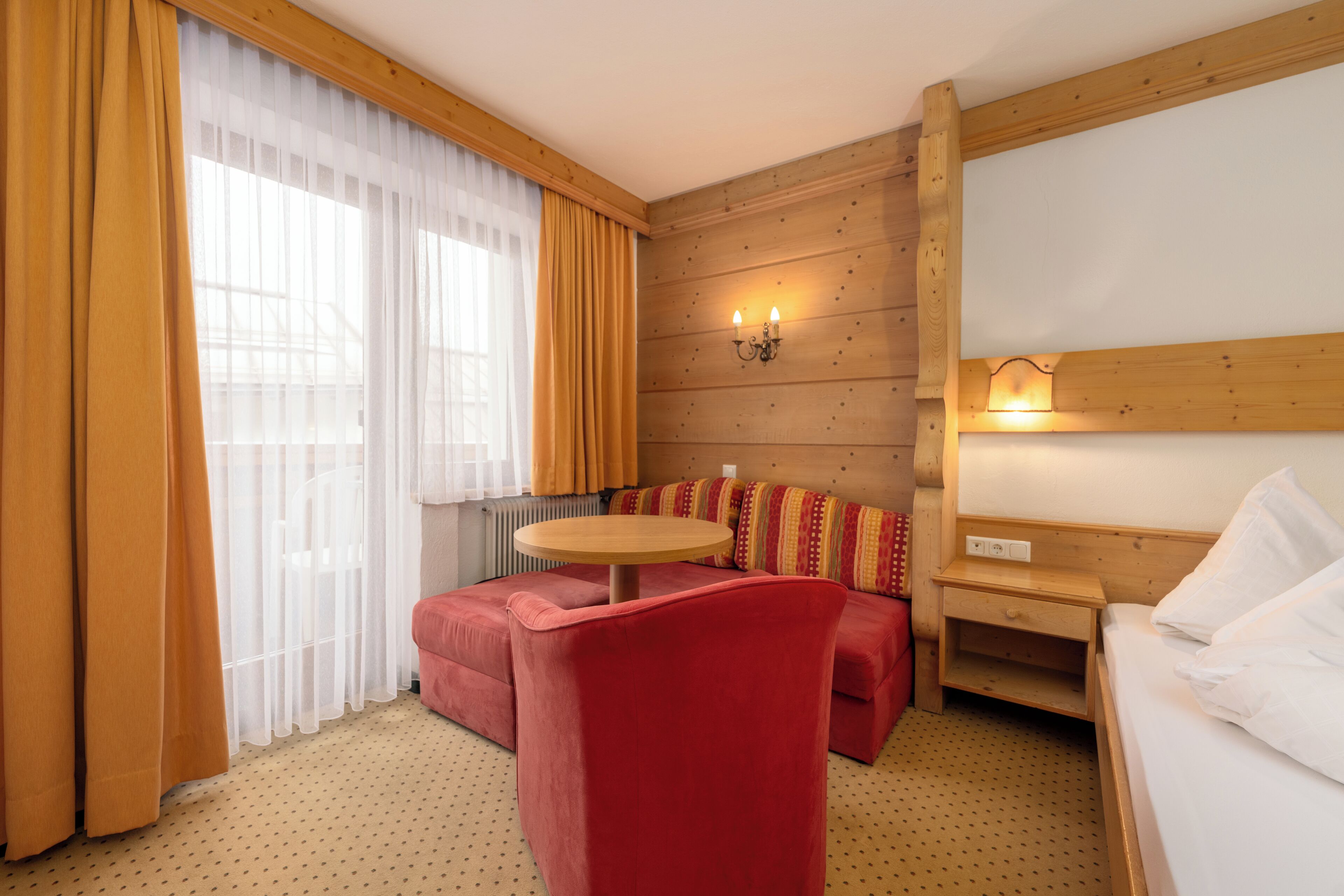 Una habitación de hotel con pared de madera, una cama, una silla roja, una mesita pequeña y un sofá.