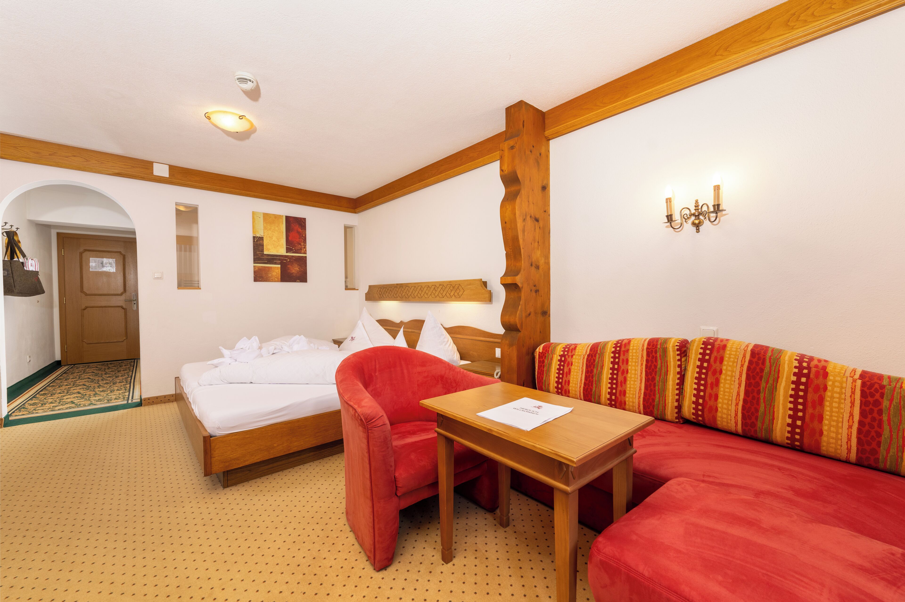 Ein Hotelzimmer mit einem Bett, einem roten Sessel, einem Holztisch und einer Couch mit einem farbenfrohen Kissen.