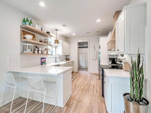 A modern kitchen with white cabinetry, a central island with bar stools, wooden flooring, and a potted plant.