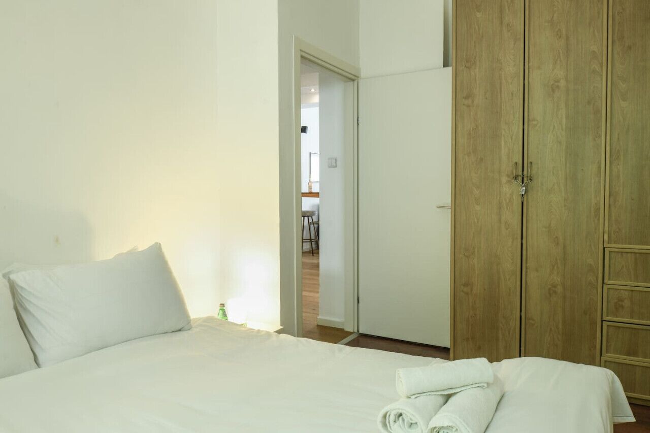 Un dormitori amb un llit, almoines blanques, un armari de fusta i una porta que dona a una altra habitació.