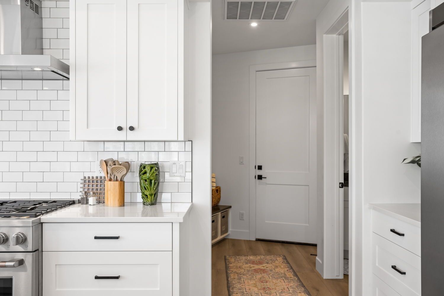 A modern kitchen with white cabinetry, a gas stove, and a built-in oven. There is a small island with a vase and kitchen utensils. A hallway with a door and a rug is visible.