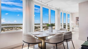 Comedor con mesa dispuesta para seis personas, ventanas amplias y vistas a un paisaje urbano.