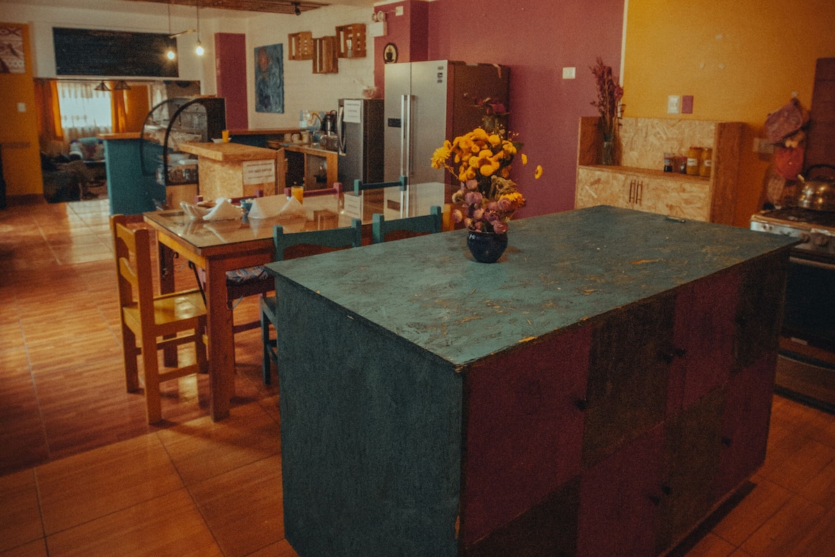 A kitchen with a central island, wooden chairs, a dining table, and hanging pendant lights.