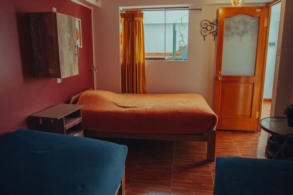 A room with two beds, a window with yellow curtains, a wooden door, and a wall-mounted shelf with books.