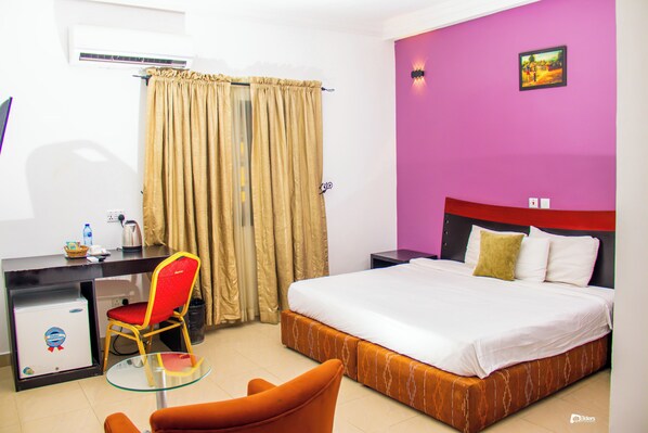 A hotel room with a bed, a desk with a chair, a small table, and a window with curtains.