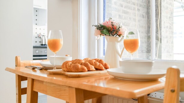 Esmorzar amb croissants, dues flaites d'espréms d'aranja i un ram de flors sobre una taula de fusta vora una finestra.