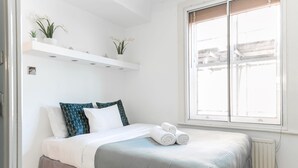 Un llit ben fet amb una falda gris, almoinades blaves i blanques estampades i tovalloles blanques arremolinades damunt del llit. Hi ha dues estanteries blanques per sobre del llit amb plantes en testos.