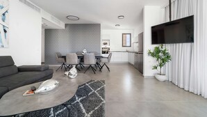 A modern living room with a grey sofa, a coffee table, and a dining area with chairs and a table. A flat-screen TV is mounted on the wall, and there is a potted plant in the corner. - Tranquil 2Bdr Oasis In The Heart Of Jerusalem (Jerusalem)