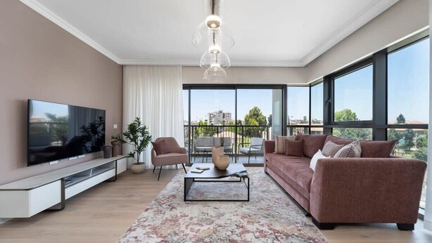 Un salon moderne doté d’un téléviseur à écran plat, d’un canapé, d’une table basse et d’un balcon offrant une vue sur les immeubles et les arbres.