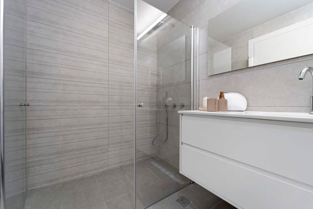 A modern bathroom with a glass shower enclosure, a white vanity with a mirror, and a tiled floor.