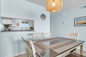 A dining area with a glass table, beige chairs, a woven light fixture, a clock, and a framed picture on the wall. - Beach Birds: Oceanfront Condo with Excellent Views (Oak Island)