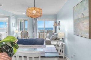 A room with a dining table, chairs, a sofa, a hanging light fixture, and a view of the sea through large windows. - Beach Birds: Oceanfront Condo with Excellent Views (Oak Island)