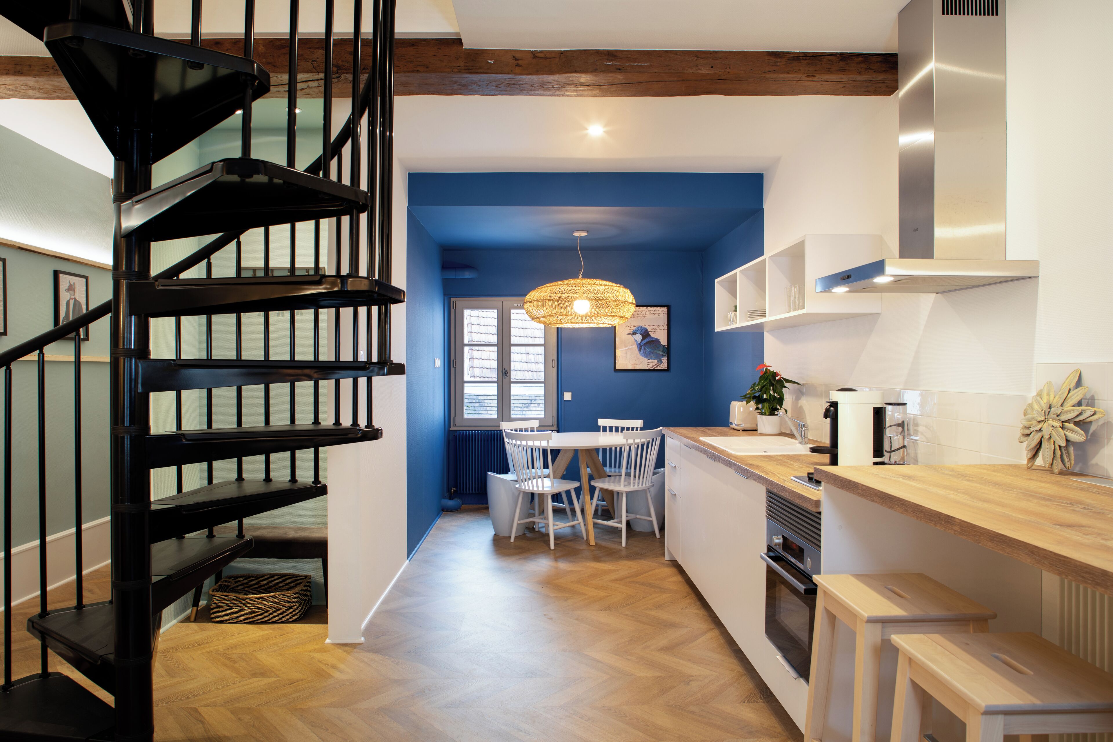 Een moderne keuken met een houten trap, een eethoek met tafel en stoelen, en een blauwe accentmuur.