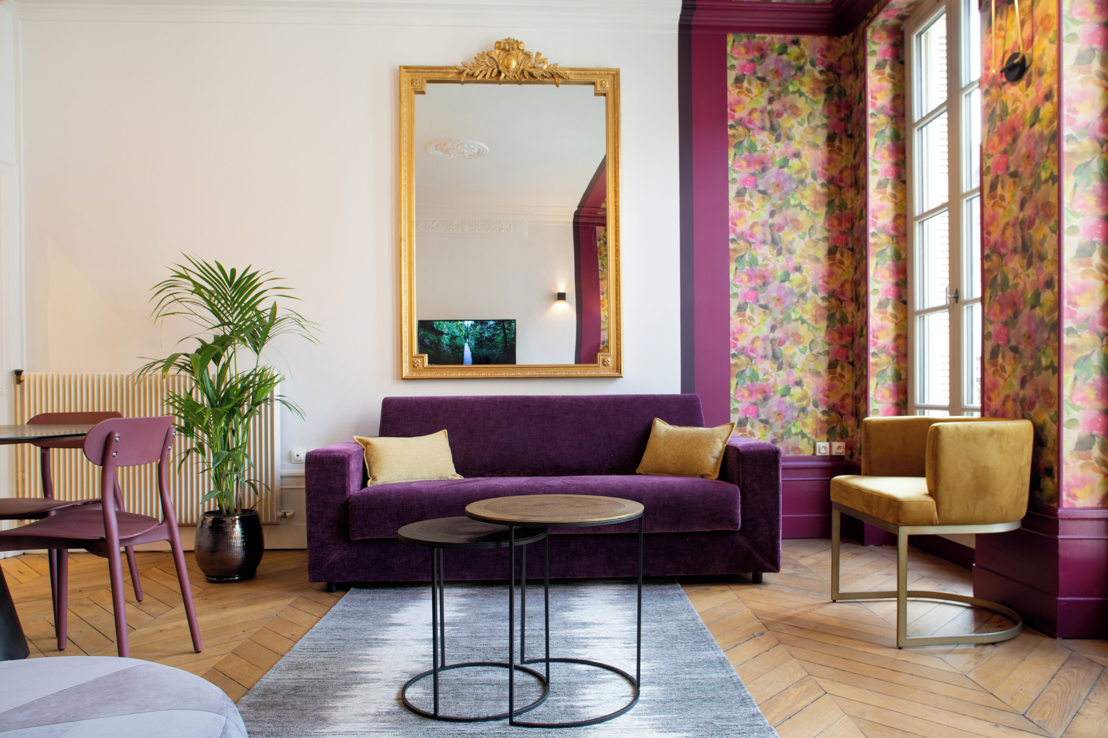 Una sala de estar con un sofá morado, dos mesas redondas, un espejo grande y una planta en maceta.