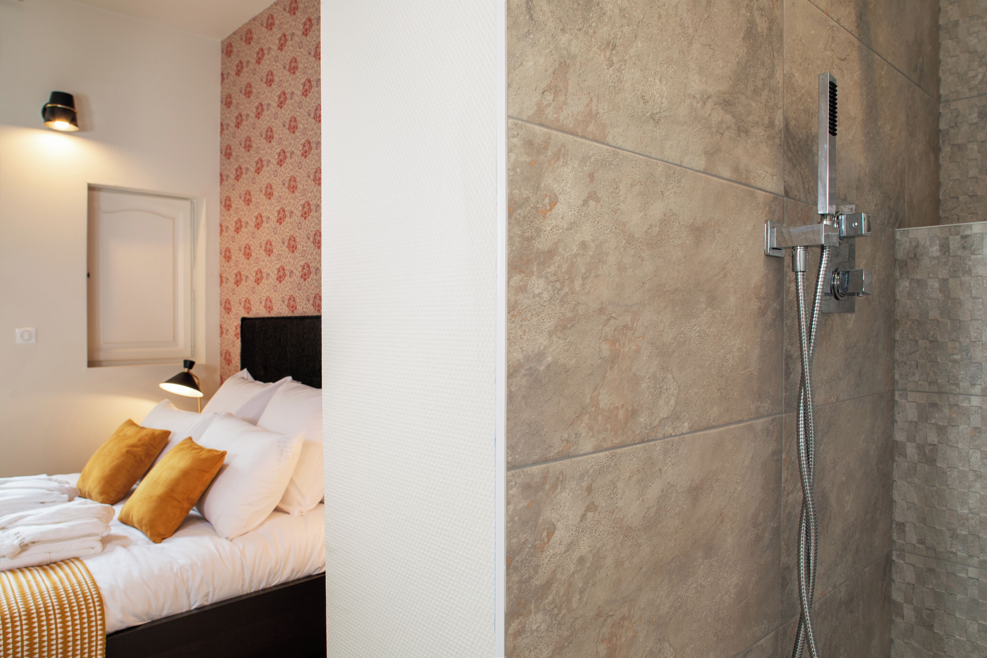 Habitación de hotel con cama, mesita de noche, lámpara y zona de ducha con cabezal de ducha manual.