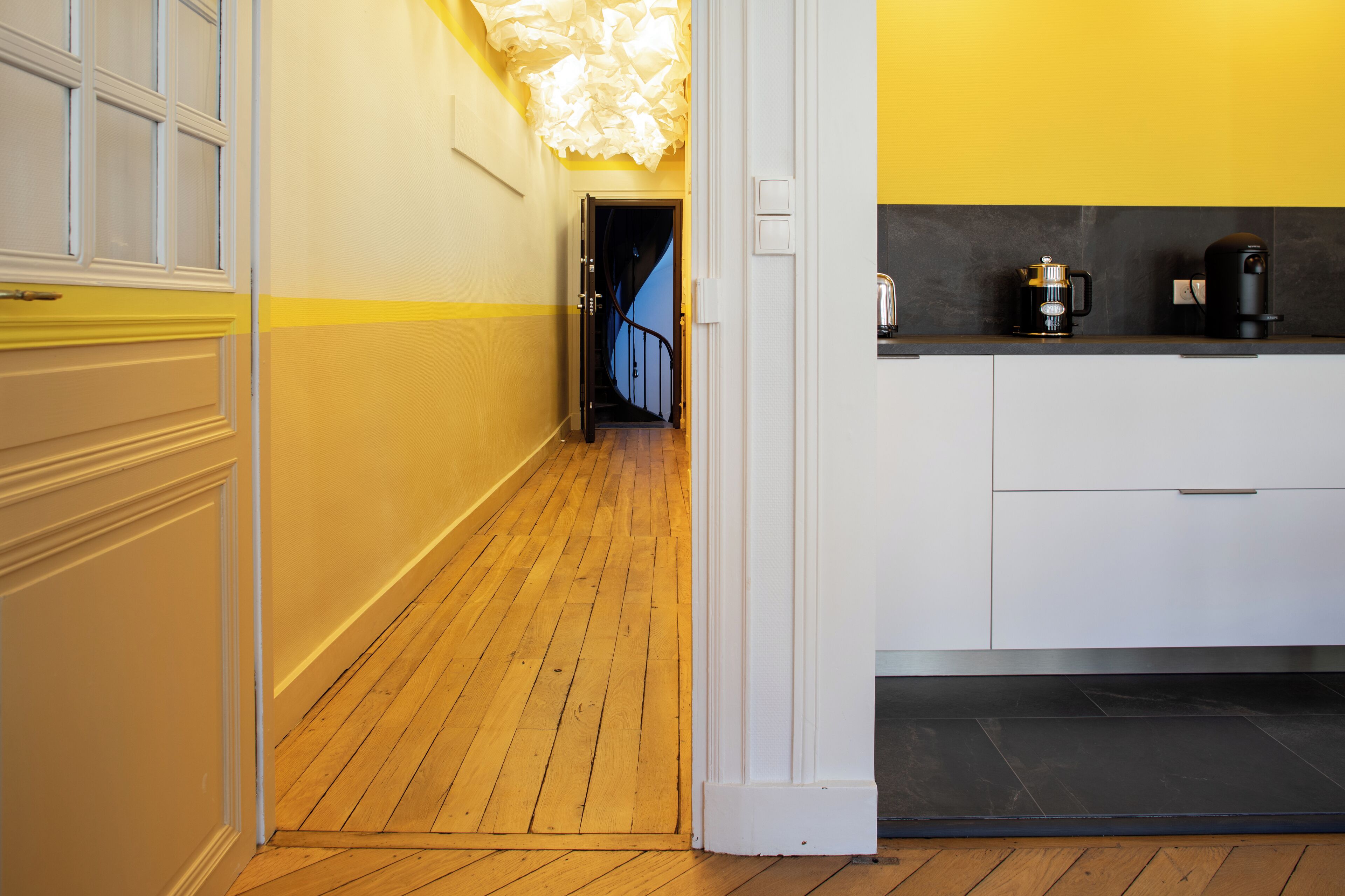 Un pasillo con piso de madera, paredes blancas y una pared de acento amarilla. Se ve una escalera que conduce a otro nivel y una zona de cocina con una cafetera y un hervidor.