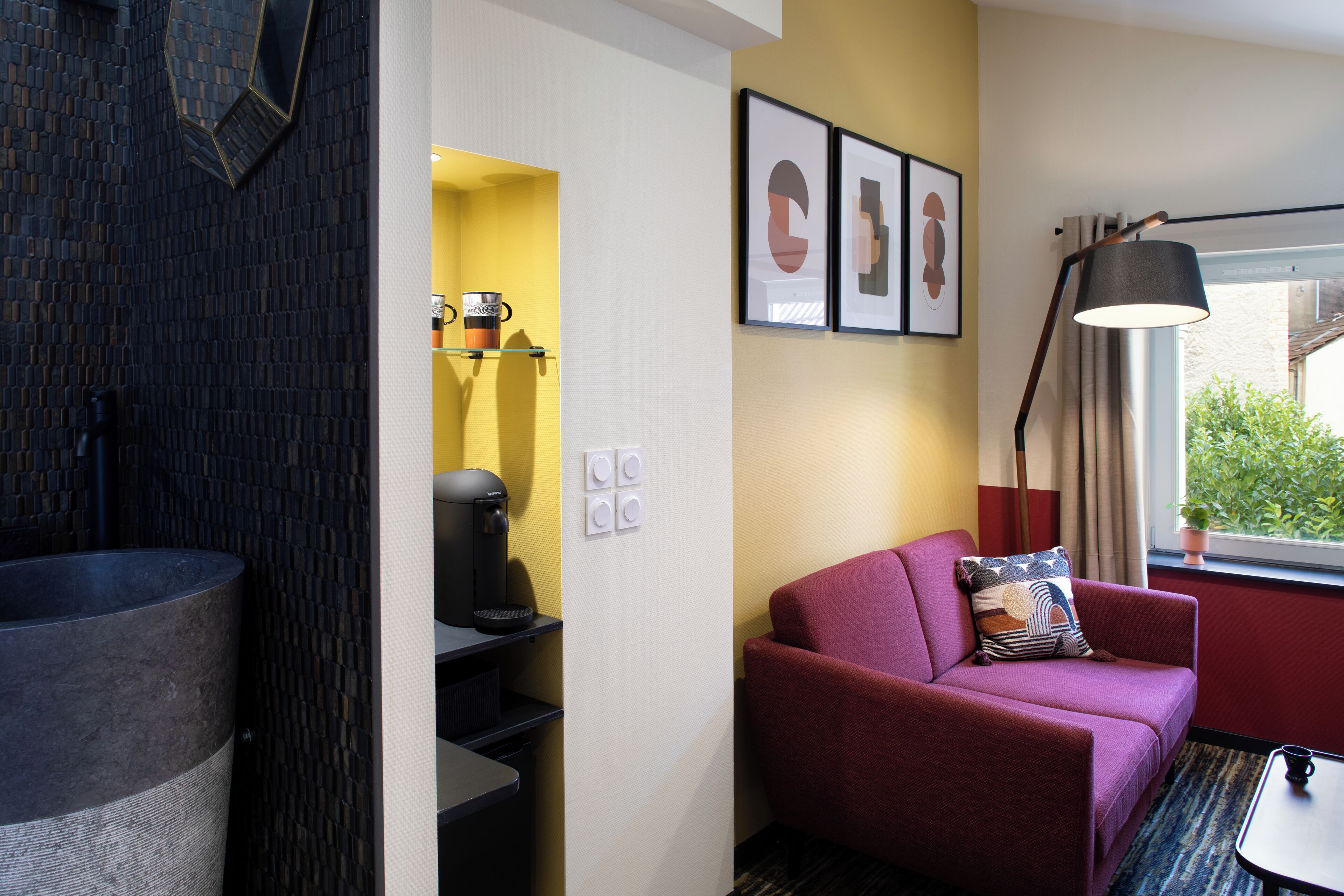 Une chambre d’hôtel moderne avec un objet cylindrique noir, un canapé rouge orné d’un coussin à motifs, une petite table et une fenêtre donnant sur un paysage verdoyant.