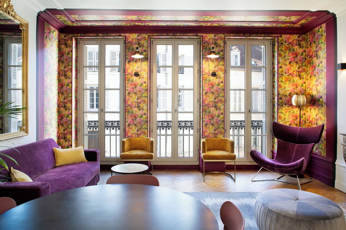 Une pièce avec un canapé violet, deux fauteuils jaunes, une table ronde et une chaise violette.