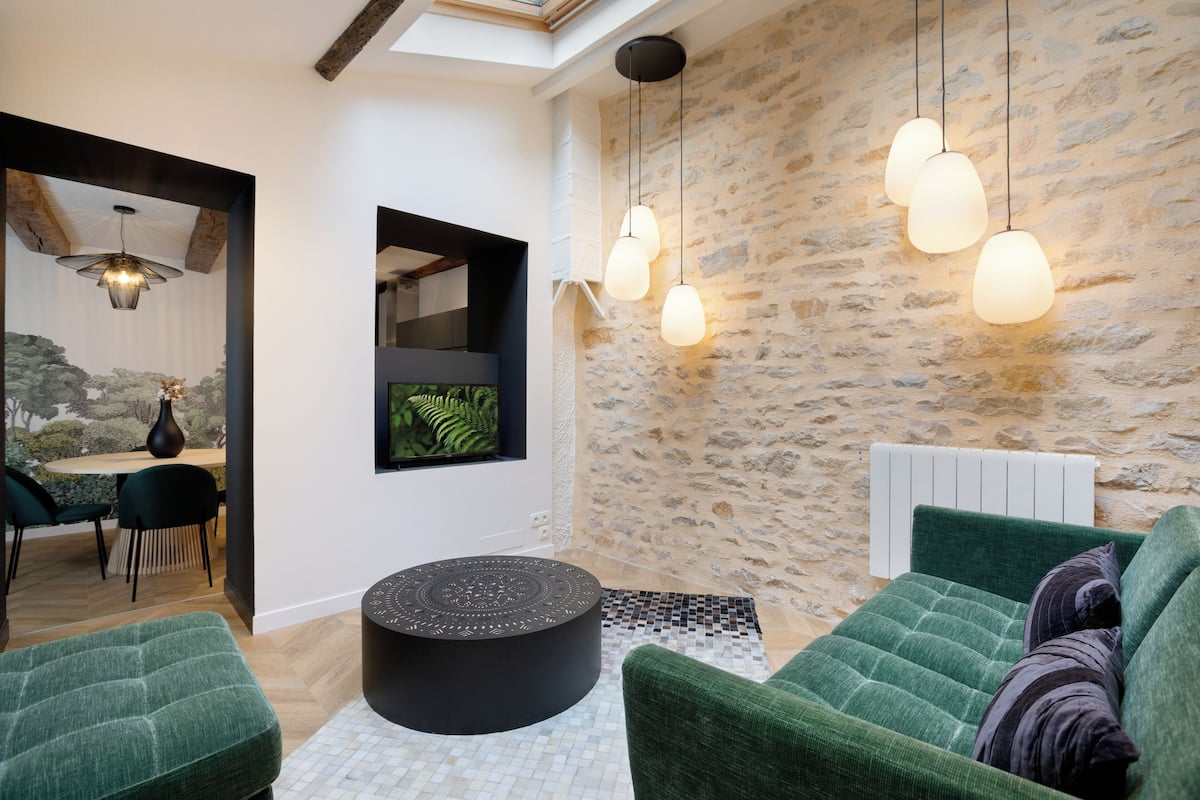 Un salon moderne avec un mur en pierre, des canapés verts à motifs, un pouf noir et des luminaires suspendus.