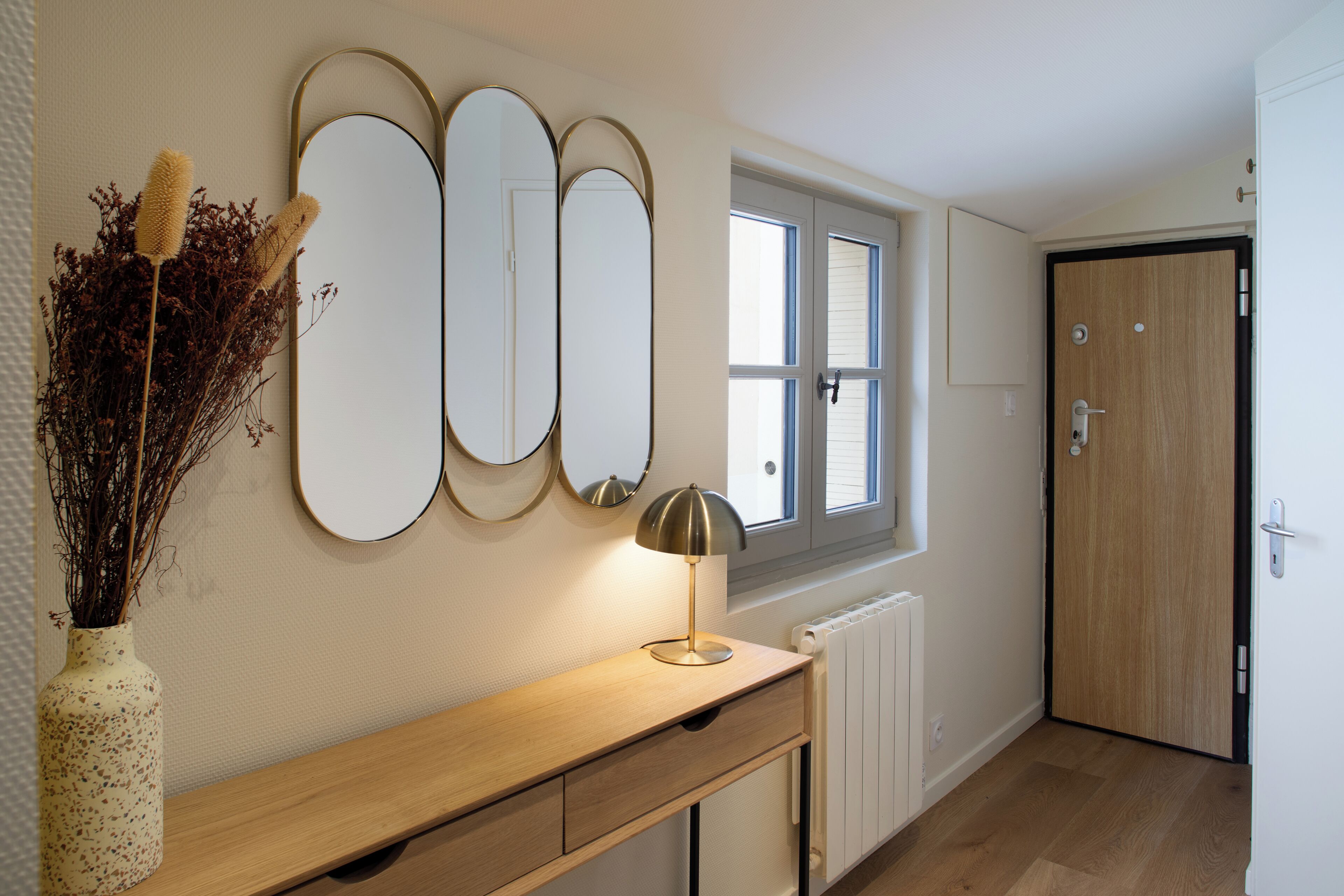 A hallway with a wooden console table, a vase with dried plants, three wall mirrors, a window, and a door.