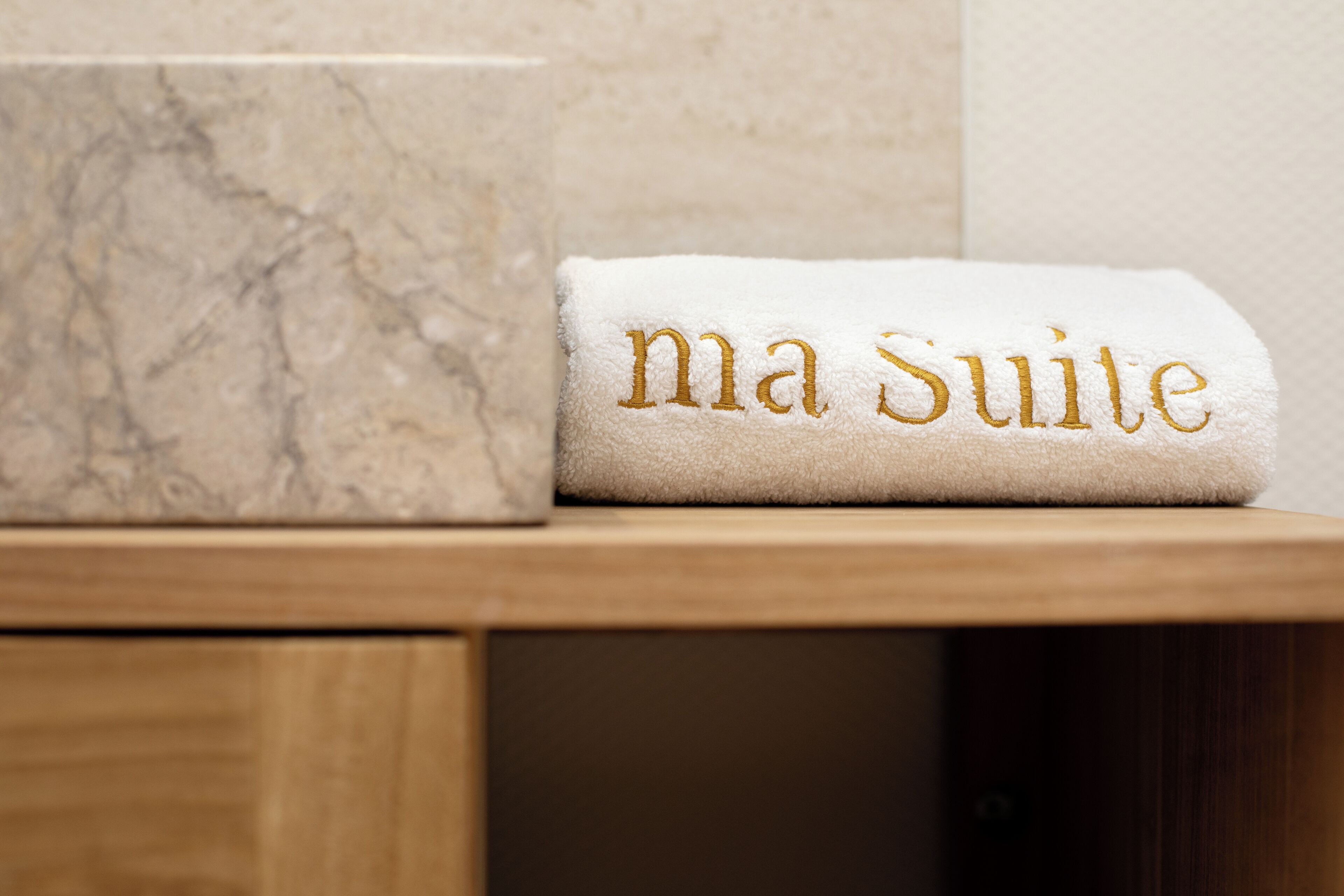 Et hvidt håndklæde med "ma Suite" broderet i guld, placeret på en træoverflade ved siden af en marmorlignende genstand.