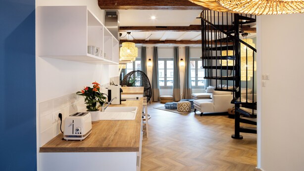 Une cuisine moderne avec un plan de travail en bois, des armoires blanches et un grille-pain encastré. Le salon est agrémenté d’un fauteuil en osier et d’un canapé blanc. Un escalier en colimaçon noir mène à l’étage supérieur.