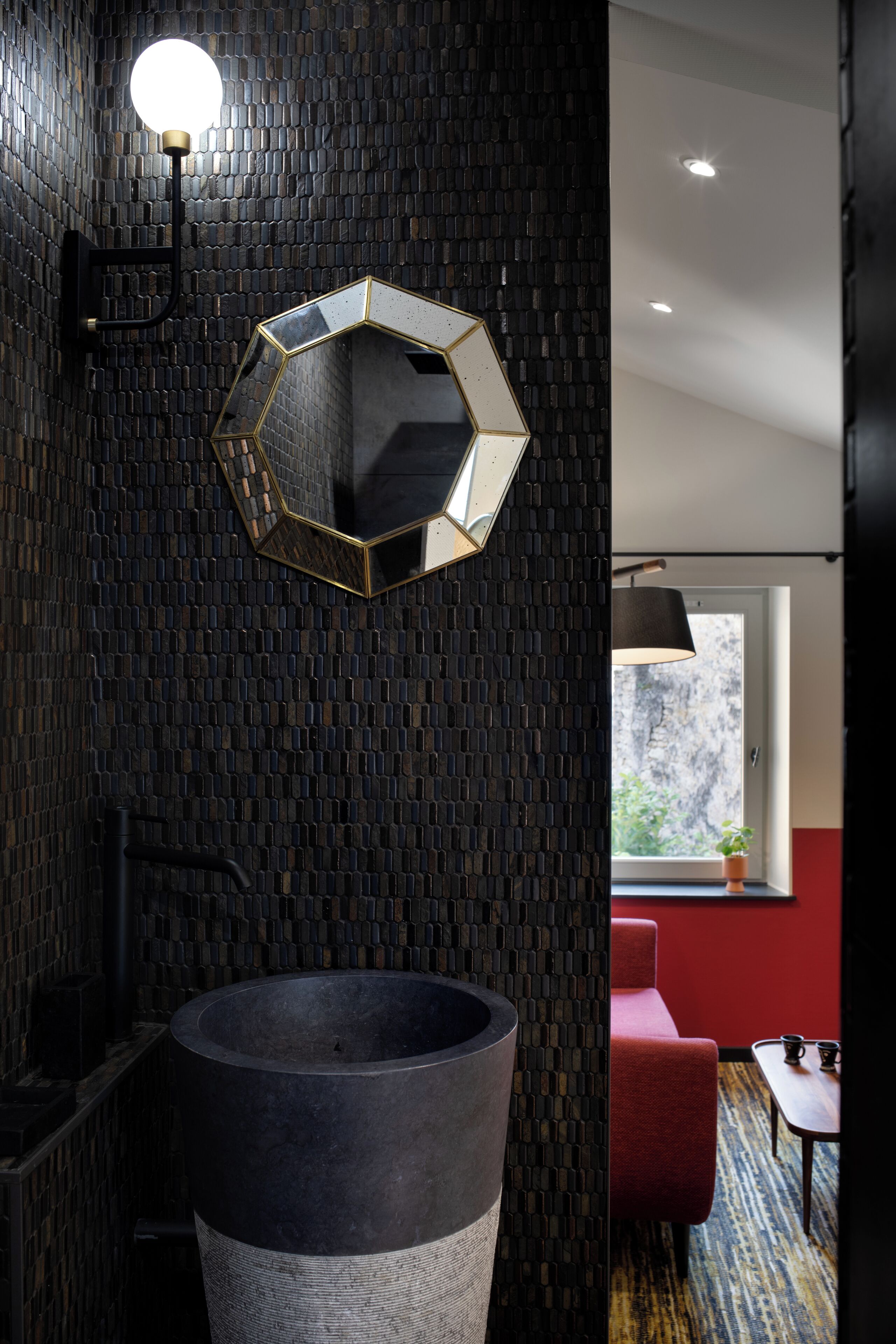 Un bagno con uno specchio esagonale, un lavandino nero di grandi dimensioni e un divano rosso nella stanza adiacente.