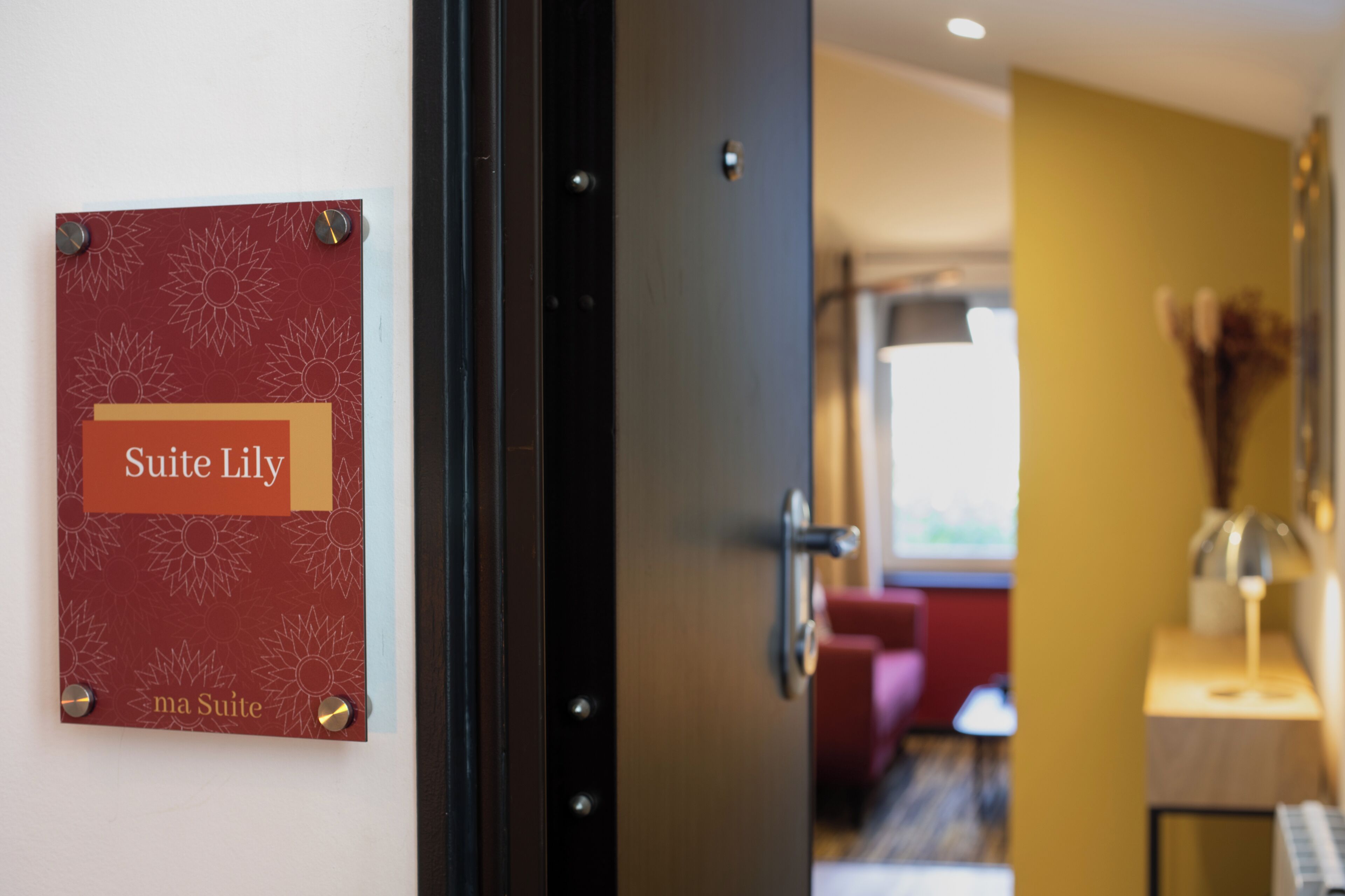 A hotel room entrance with a sign reading "Suite Lily" and a door leading to a room with a red sofa and a lamp on a bedside table.