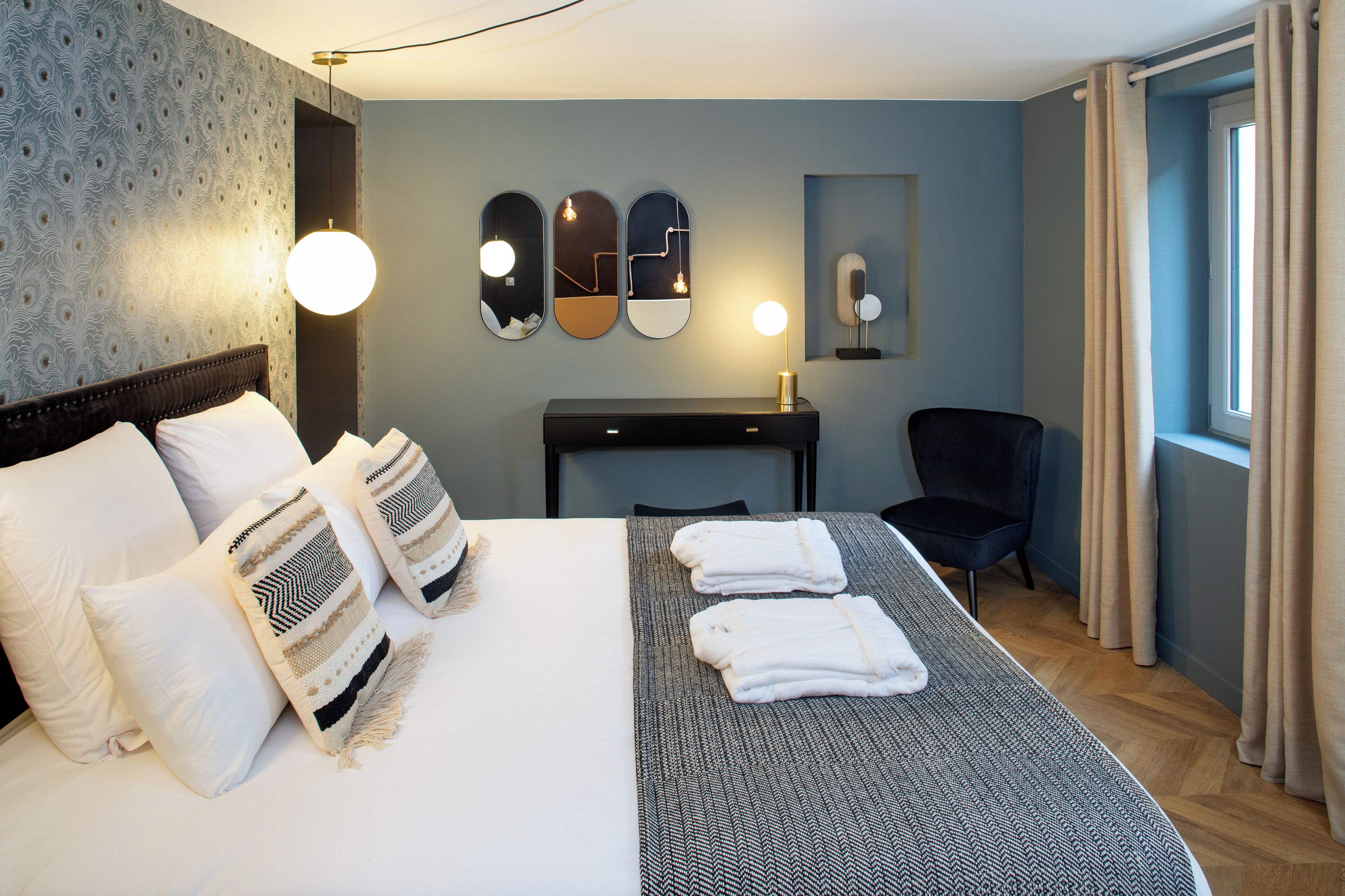 A hotel room with a neatly made bed, a desk with a lamp, a chair, and a large window with curtains.