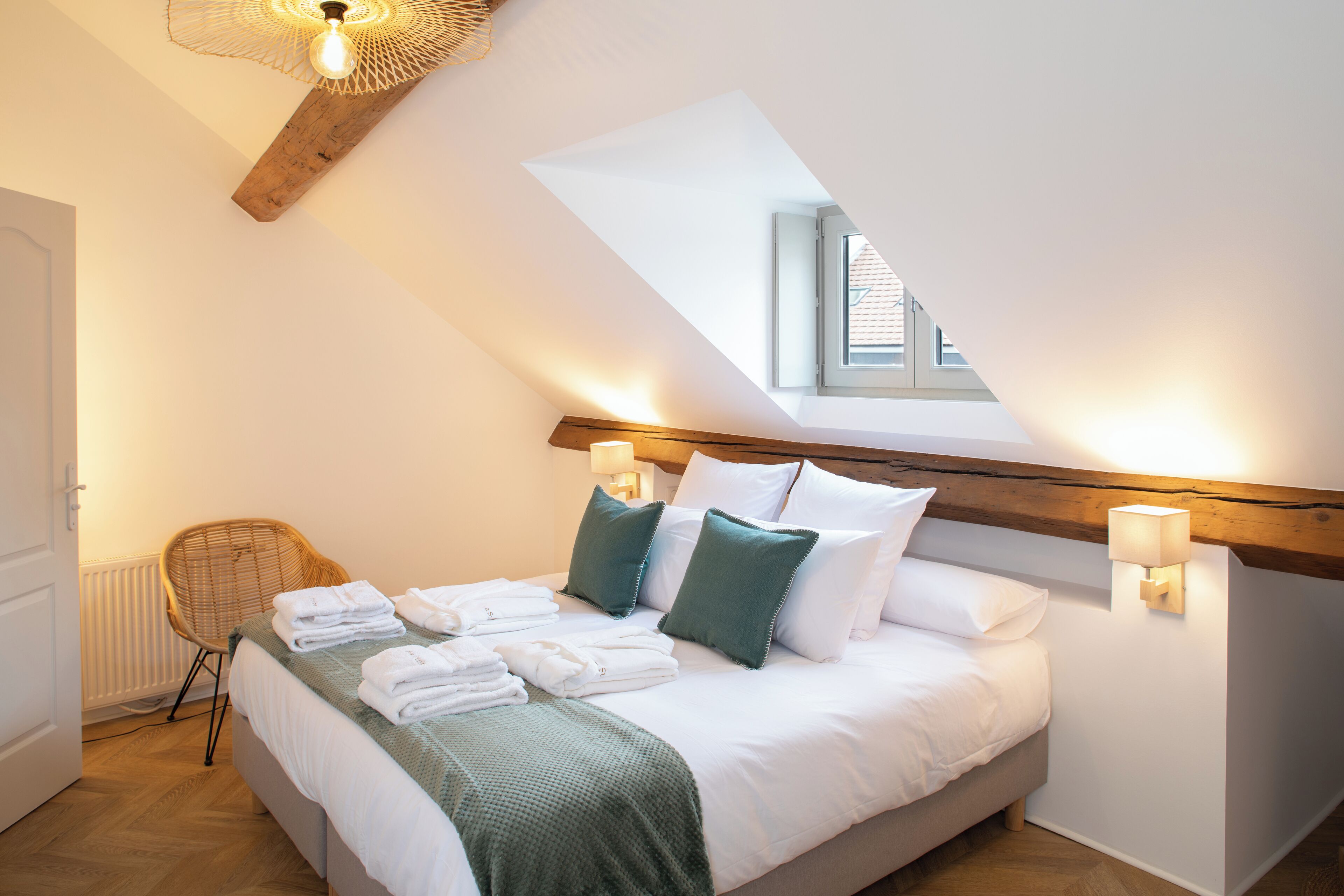 Un dormitorio con techo inclinado, tragaluz, cama con ropa de cama blanca, almohadas verdes y una silla.