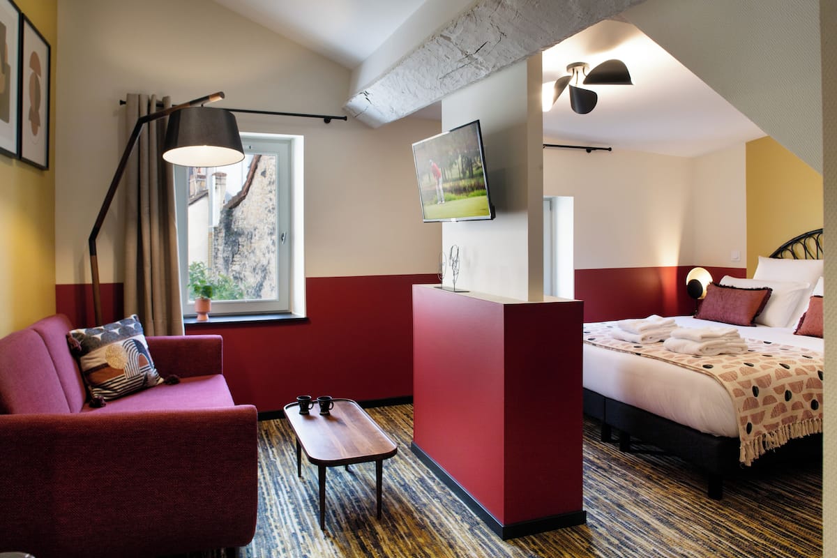Une chambre d’hôtel aux couleurs rouge et blanc, avec un lit recouvert d’une couverture à motifs, un bureau avec une lampe, un téléviseur fixé au mur et une fenêtre donnant sur un bâtiment à l’extérieur.