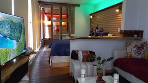 Habitación de hotel con un televisor grande, una cama con una colcha azul, un sofá con una manta roja y una mesita con un jarrón y velas.