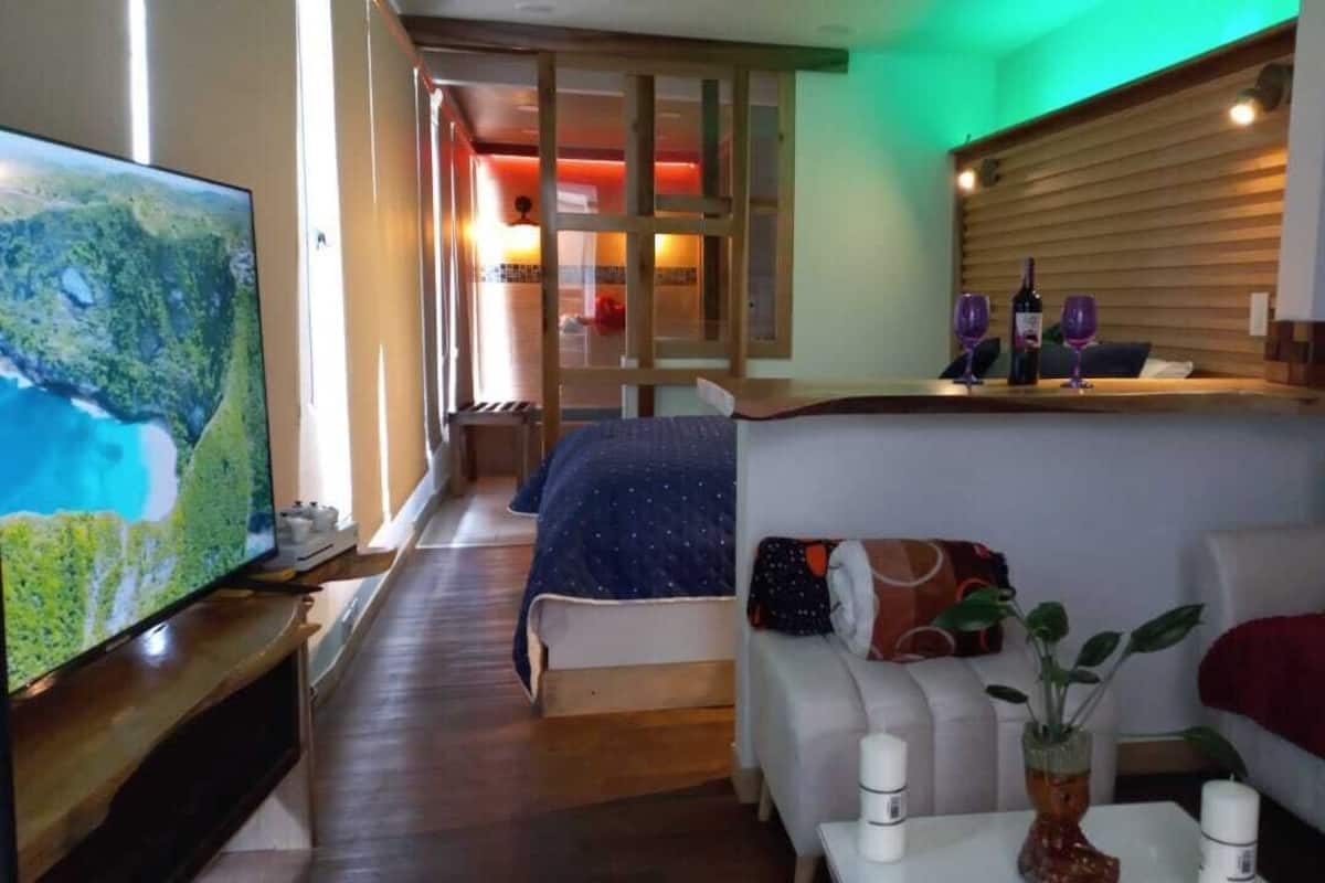 A hotel room with a large TV, a bed with a blue blanket, a sofa with a red blanket, and a small table with a vase and candles.