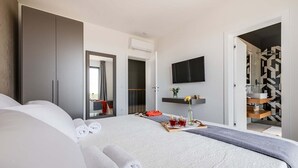 Ein modernes Hotelzimmer mit einem großen Bett, einem Flachbildfernseher, einem kleinen Tisch mit einer Getränderunde und einem durch eine offene Tür einsehbaren Badezimmer.