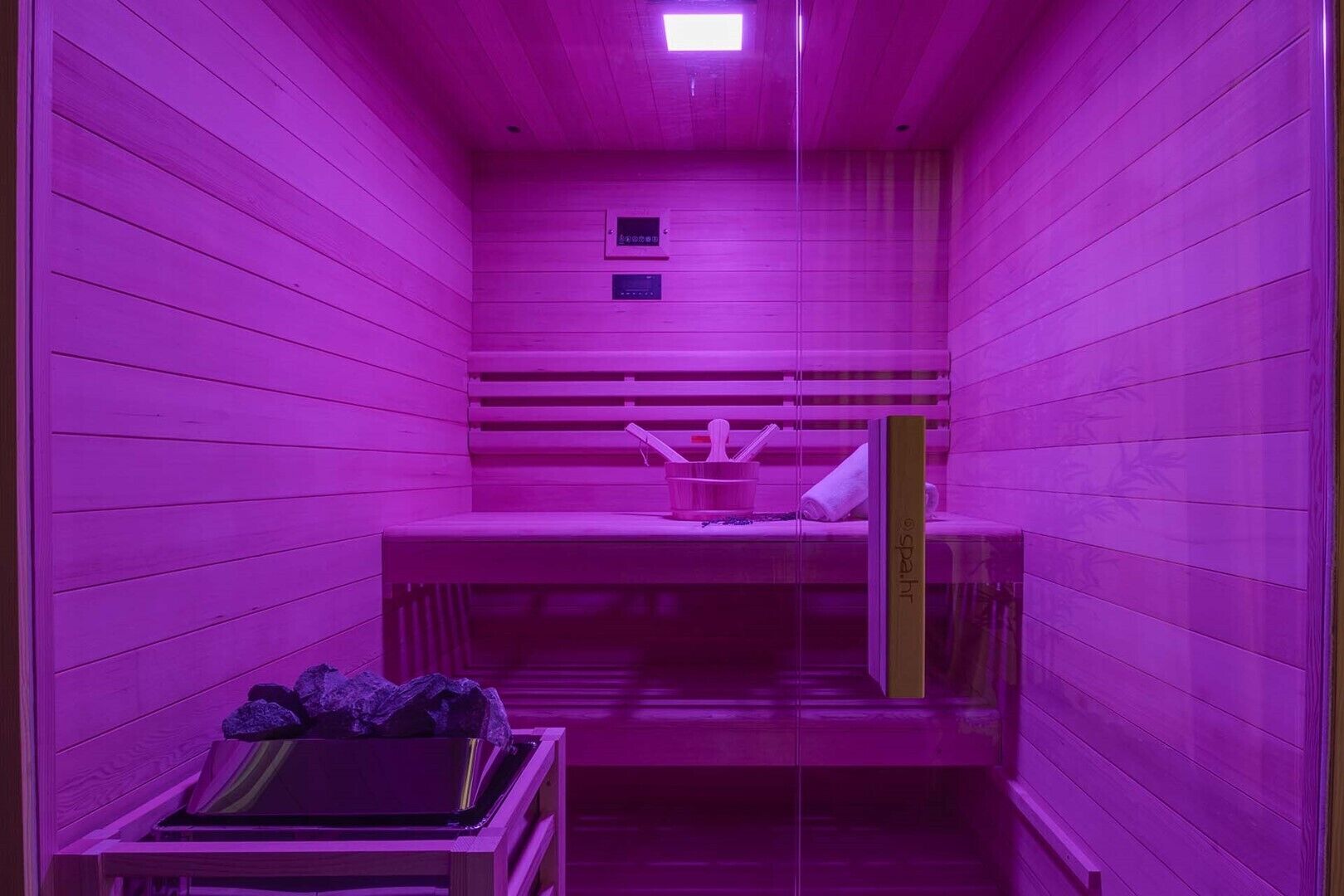 A sauna room with wooden walls and benches, a bench with a towel and a bowl, and a small window with controls.