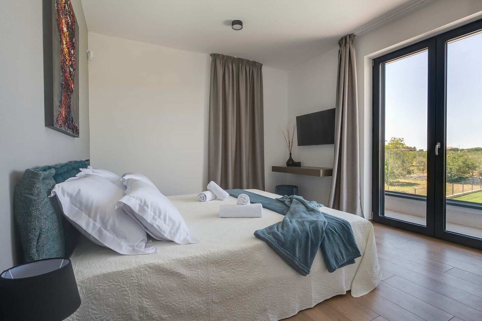 A modern bedroom with a large bed, a flat-screen TV, and a view of the outdoors.