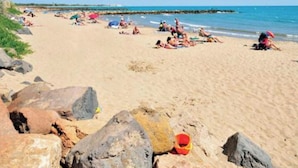 A beach with people sunbathing and swimming, large rocks in the foreground, and a clear blue sky.