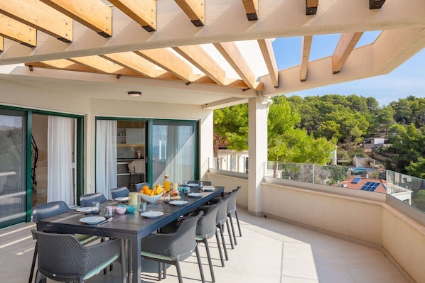 A balcony with a dining table set for four, a glass railing, and a view of a forested hillside. - Luxury Villa Leisure Seaside Brac with Pool (Milna)
