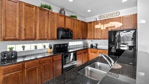 A modern kitchen with wooden cabinets, a black countertop, stainless steel sink, and a refrigerator. - Large 7 Bed In Solterra Pool, Spa Near Disney 7 Bedroom Home by RedAwning (Davenport)