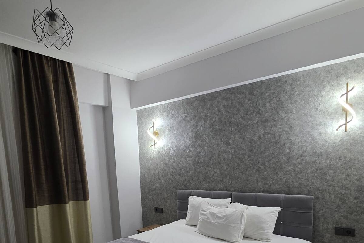 A modern bedroom with a large bed, a headboard with integrated lighting, a bedside table, and a ceiling light fixture.